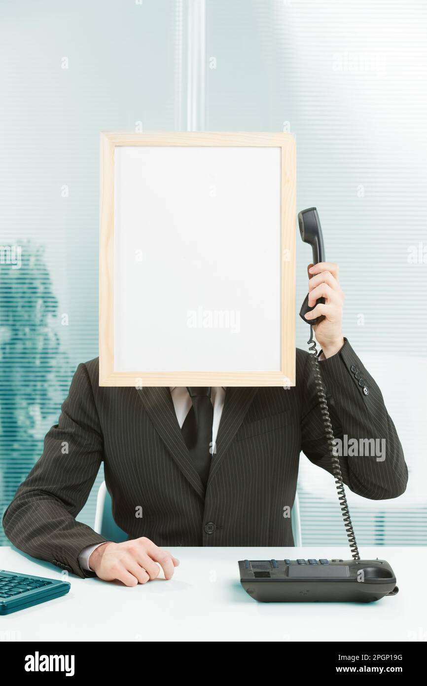 Man with framed sign head in an office answers phone. He has his arm raised with the handset in his hand, which he pulls over to the sign-head to list Stock Photo