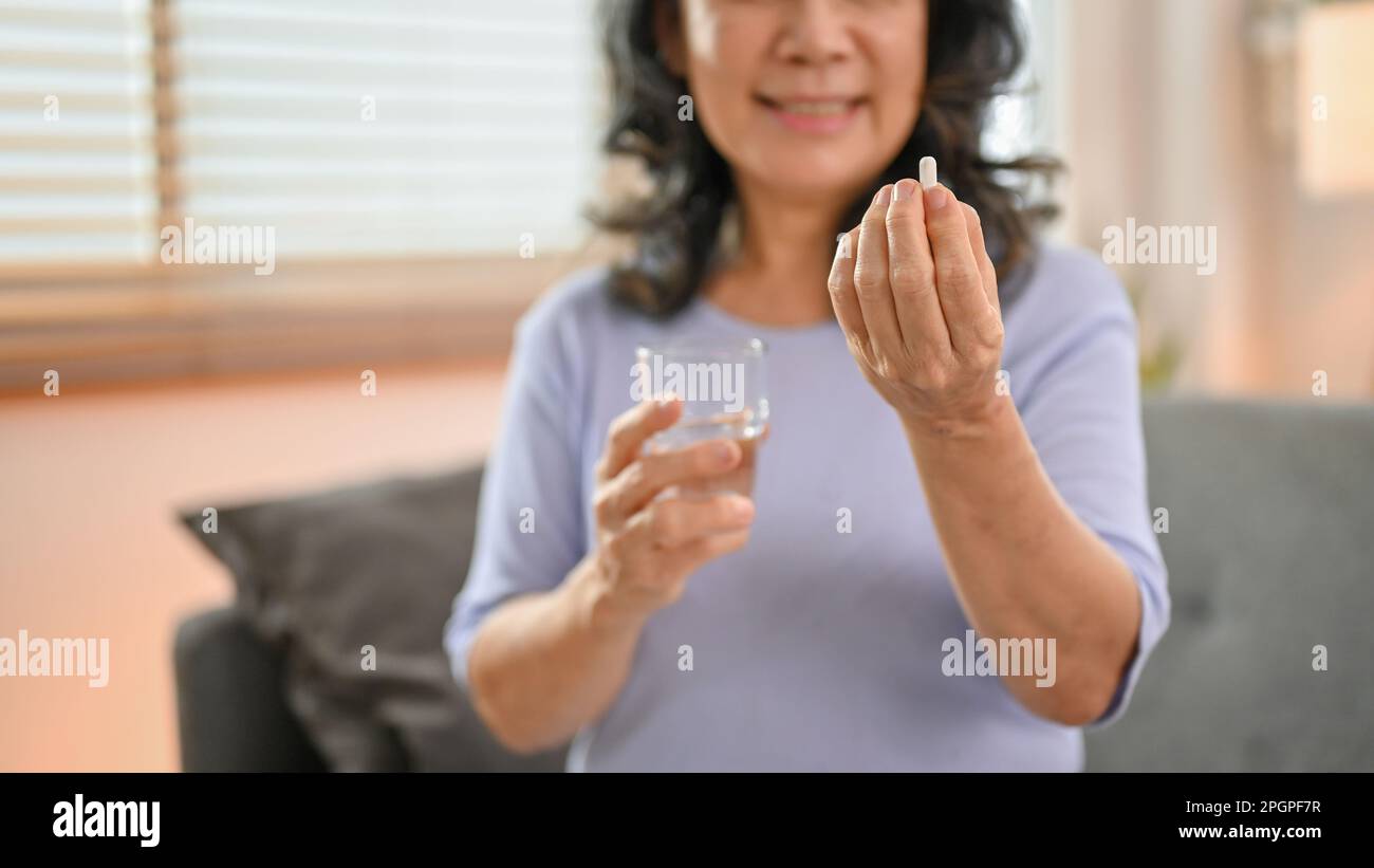 Close-up image, a happy 60-year-old retired Asian woman holding a glass of water and a pill, taking a medicine in her living room. health care, wellbe Stock Photo