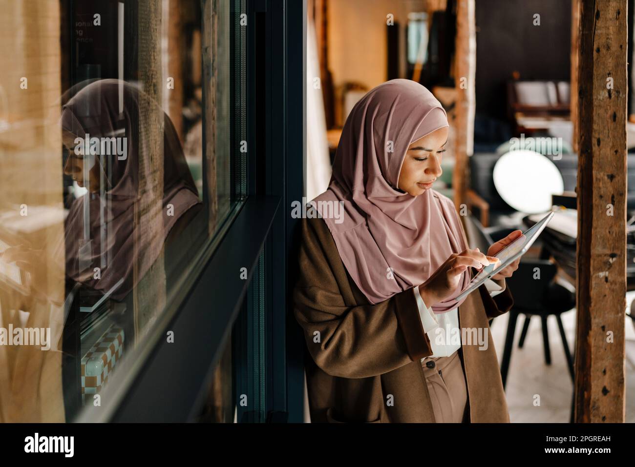 Arabian woman wearing hijab working on tablet while standing in modern ...