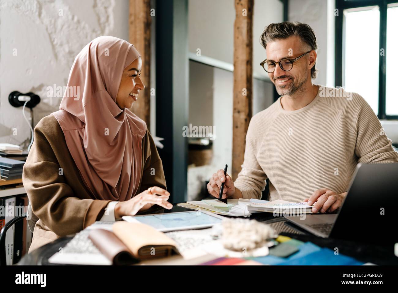 Two multinational colleagues designers smiling while working with laptop computer at workshop indoors Stock Photo