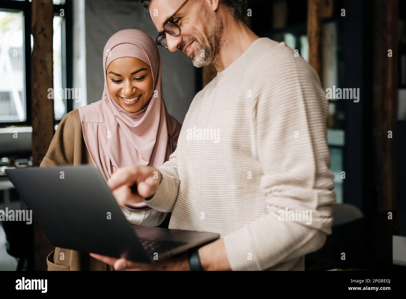 Two multinational colleagues smiling while working with laptop computer in modern office Stock Photo