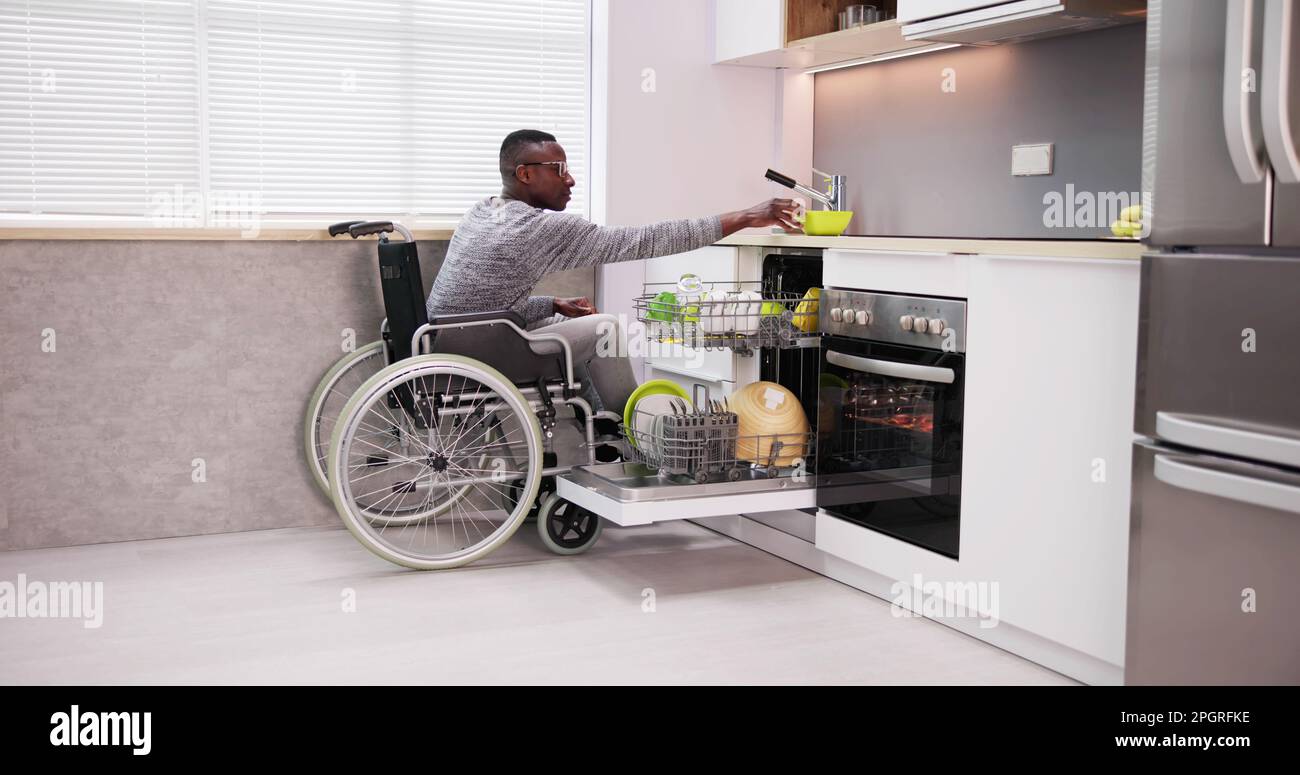 Young Man Sitting On Wheelchair Arranging Plates In Dishwasher Stock Photo