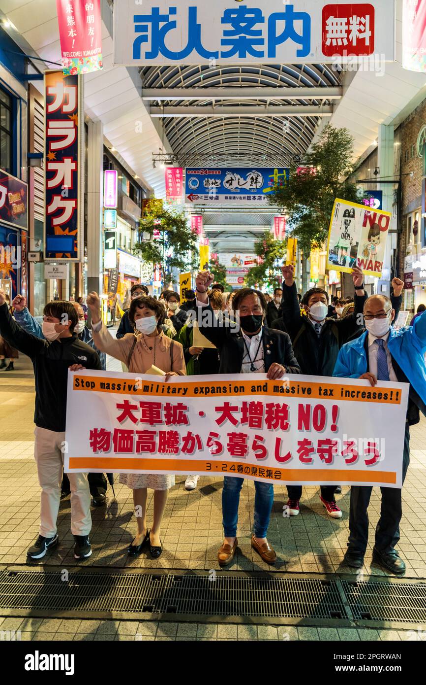 Japanese protestors marching through a shopping arcade in Kumamoto city in the evening with a banner at the front and several demonstrators holding placards. The demonstration is against increased spending on the civil defence force. Stock Photo