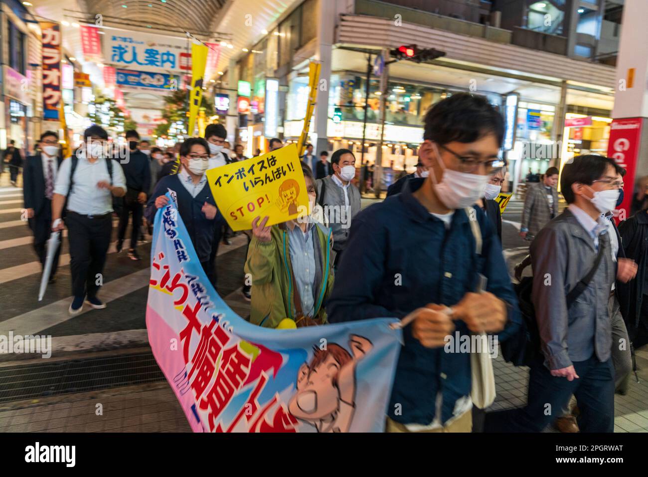 Japanese protestors marching through a shopping arcade in Kumamoto city in the evening with a banner at the front and several demonstrators holding placards. The demonstration is against increased spending on the civil defence force. Stock Photo