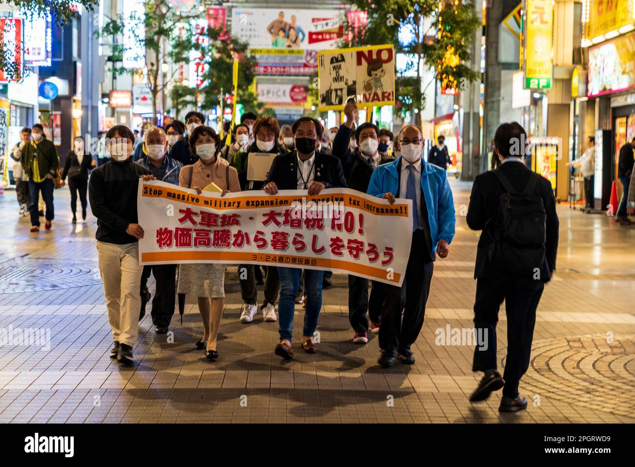 Japanese protestors marching through a shopping arcade in Kumamoto city in the evening with a banner at the front and several demonstrators holding placards. The demonstration is against increased spending on the civil defence force. Stock Photo