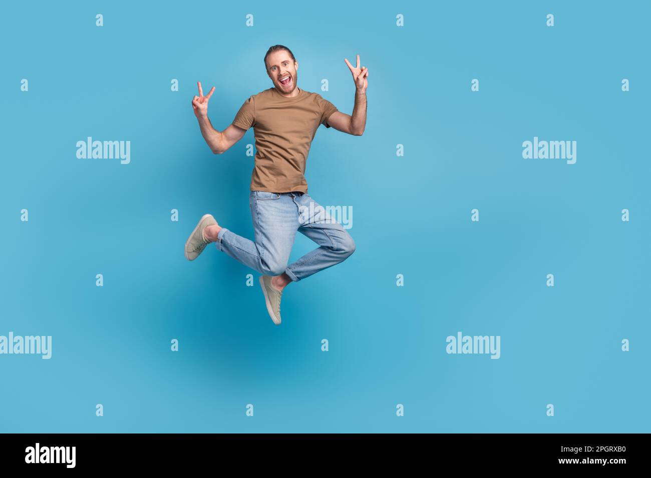 Full length photo of excited funny man wear beige t-shirt jumping high showing v-signs emtpy space isolated blue color background Stock Photo