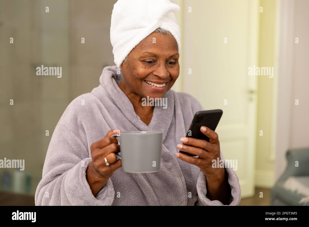 Smiling african american senior woman wearing bathrobe with coffee cup using smartphone in bathroom. Copy space, unaltered, lifestyle, bathing, drink, Stock Photo