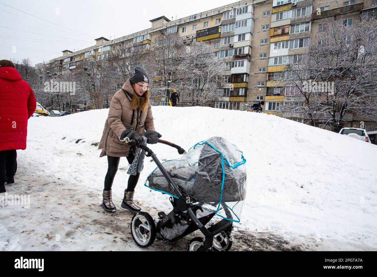 November 2022, young Ukrainian mother carefully pushes perambulator (baby carriage) on a snow hill near residential house in Kiev Stock Photo