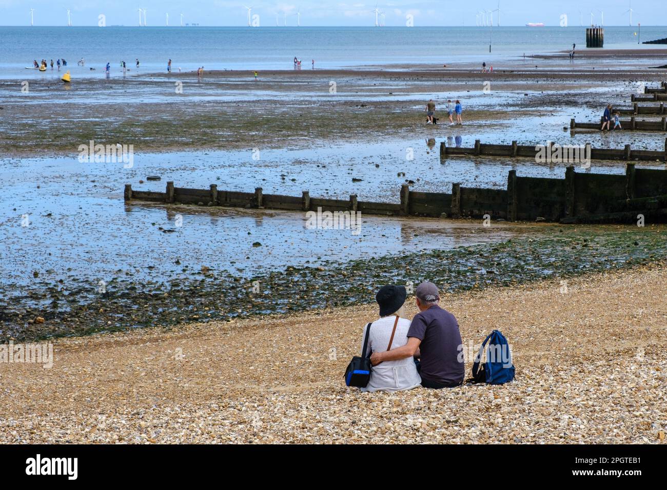 Man & woman embrace, sitting on shingle beach look out to sea & wooden groynes at Whitstable West Beach, north-east Kent coast, England, UK. Stock Photo
