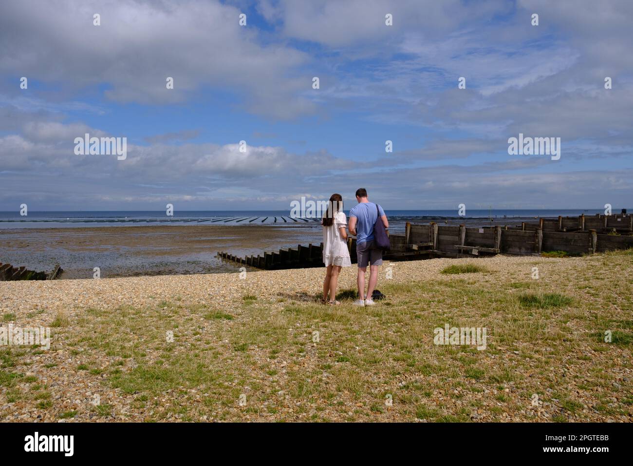Man & woman stand on shingle beach looking out to sea next to Wooden groyne. Whitstable West Beach, north-east Kent coast, England, UK. Stock Photo
