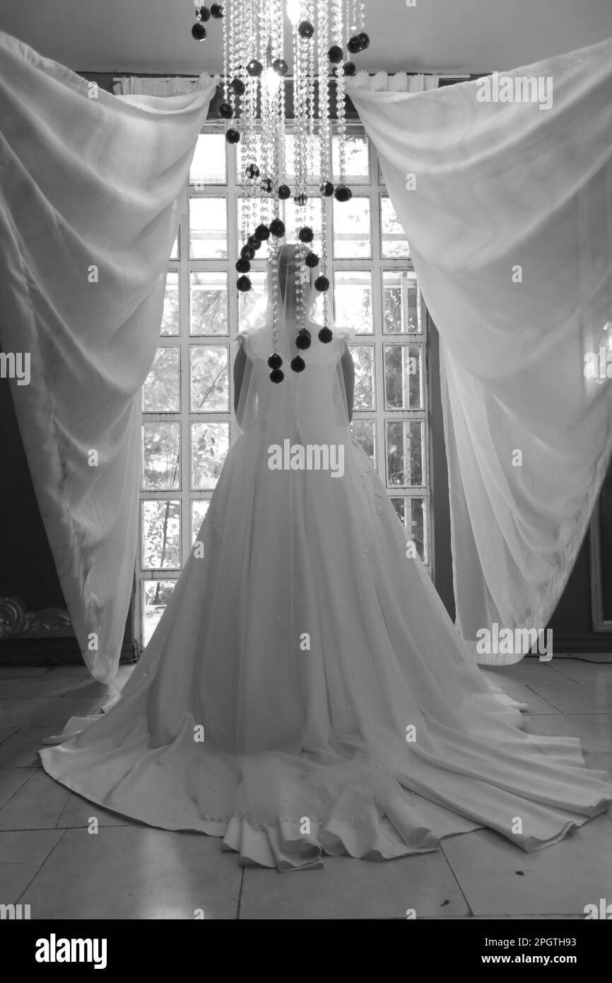 A greyscale of a happy bride standing in front of a window, wearing a white gown, with rays of sunlight streaming in through the window Stock Photo