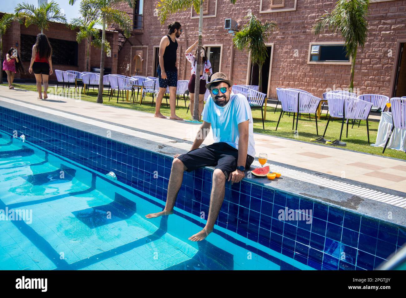 Young indian man sitting on the pool edge with feet in water and holding glass of orange juice in hot sunny day. Guy relaxing outdoors by swimming poo Stock Photo