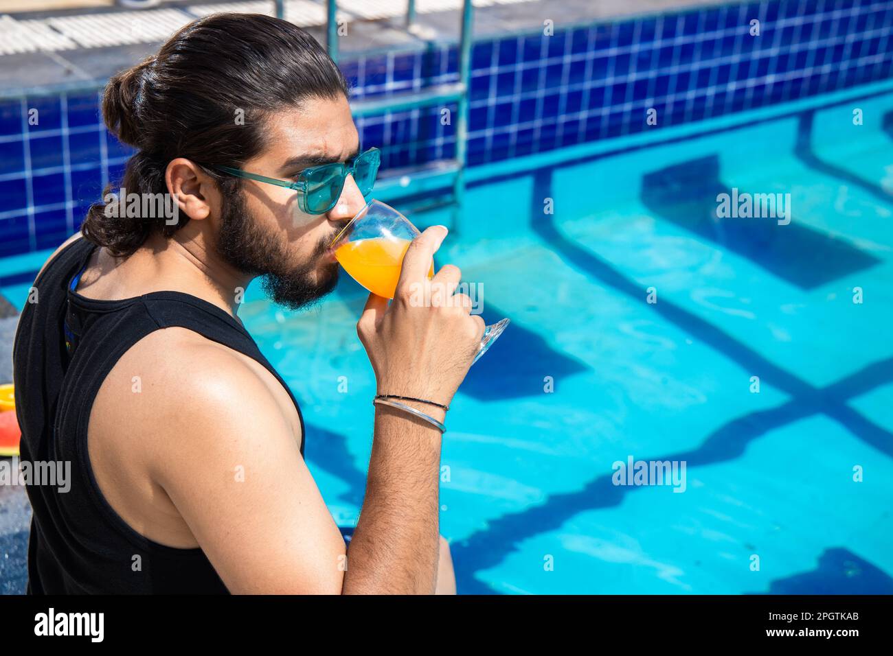 Young indian man wearing sunglasses sitting on the pool edge drinking orange juice in hot sunny day. Guy relaxing outdoors by swimming pool. Summer ho Stock Photo