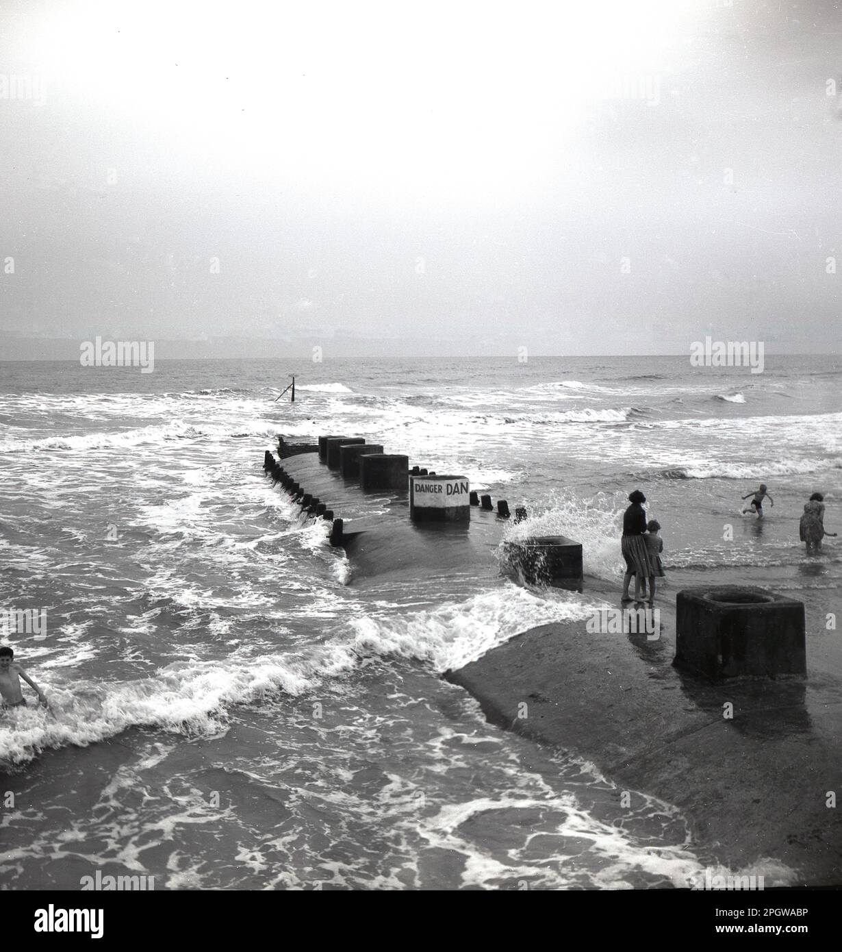 1950s, historical, children on a breakwater extending in the sea from the shore at Bridlington, North Yorkshire, England, UK......looks like a submarine emerging from beneath. Danger Sign on the concrete breakwater. Stock Photo