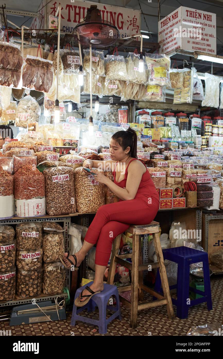 Ho-Chi-Minh City, Vietnam - Portrait of a saleswoman in a food store sitting on a bar stool engaging with her cell phone in a indoor market Stock Photo
