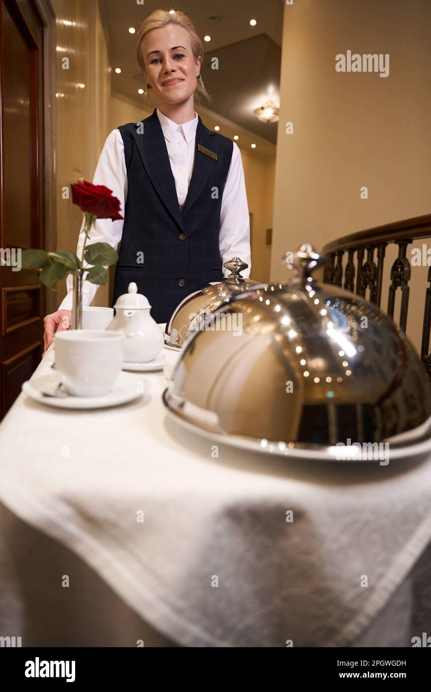 Smiling young woman on cart carrying hot food down hotel corridor