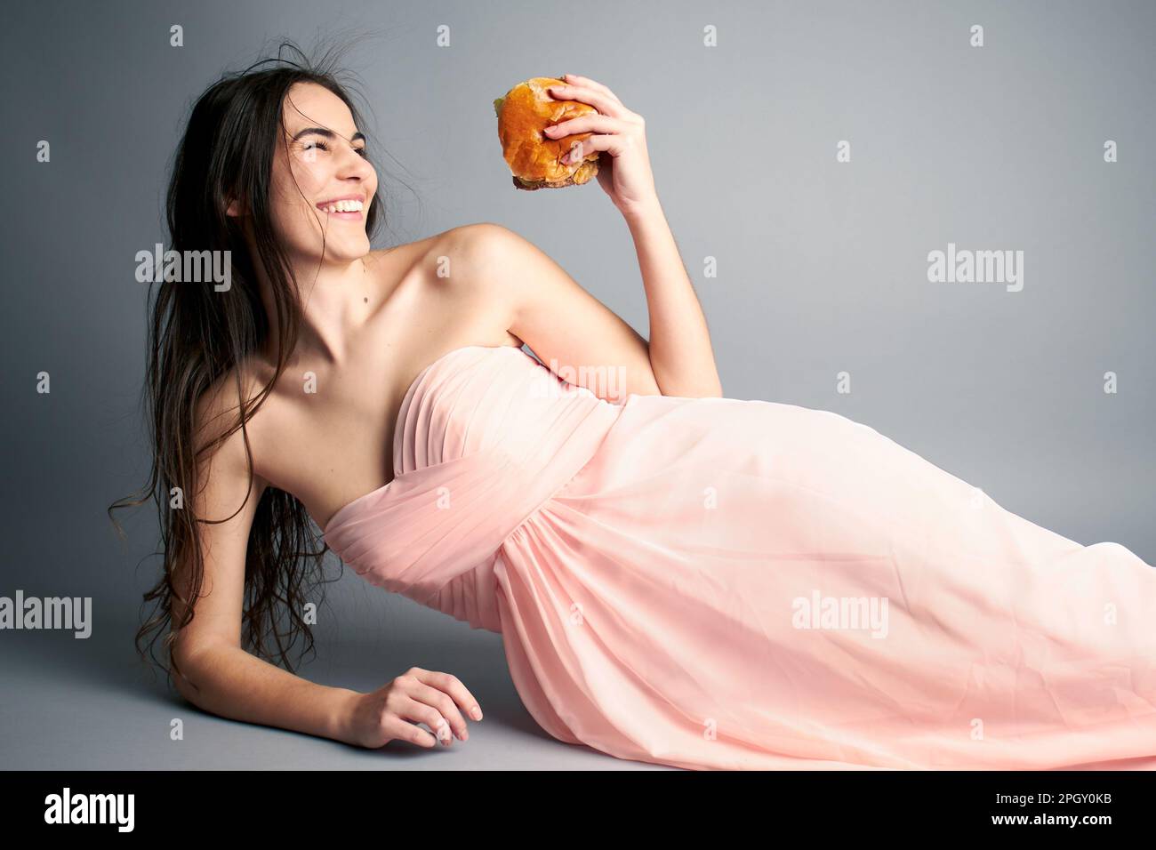 Woman in pink prom ballgown dress laying down on seamless grey paper background holding a hamburger Stock Photo