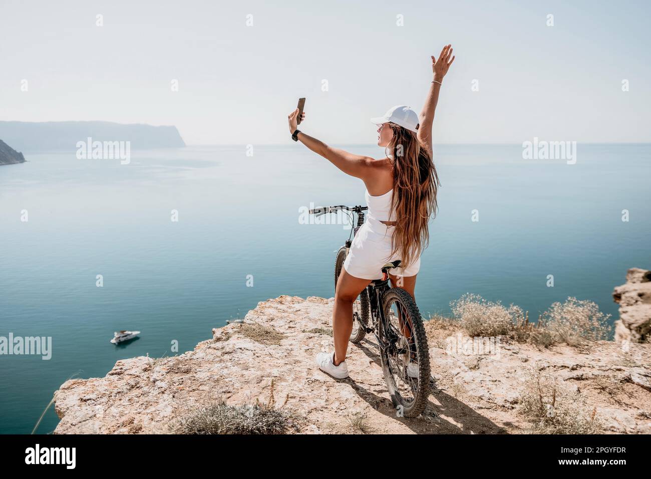 A woman cyclist on a mountain bike looking at the landscape of mountains and sea. Rear view of cyclist woman standing in front to the sea with Stock Photo