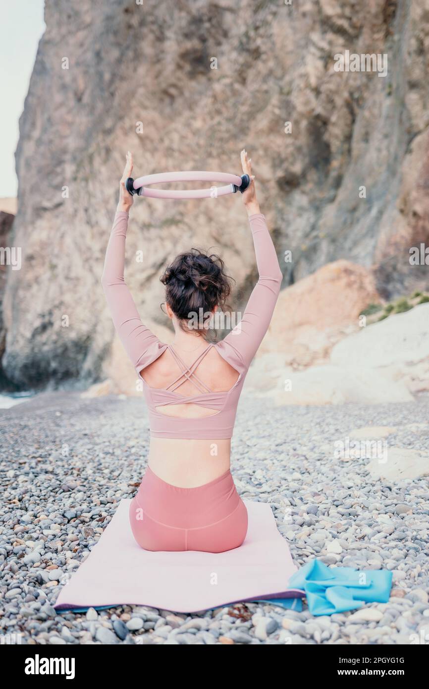 Woman sea pilates. Sporty happy middle aged woman practicing fitness on beach near sea, smiling active female training with ring on yoga mat outside Stock Photo