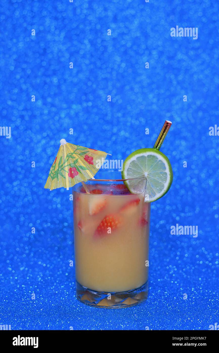 A cold, tropical, orangey, yellow coloured drink in a glass with strawberries, lime and parasol on a vibrant, bright, sparkly blue background Stock Photo