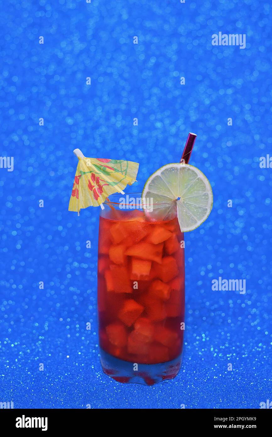 A cold, tropical, red coloured drink in a glass with orange fruit chunks, lime and parasol on a vibrant, bright, sparkly blue background Stock Photo