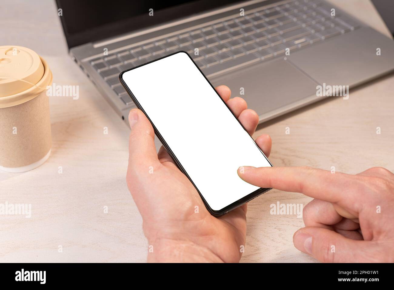Hand holding mobile phone mockup, finger clicking, touching smartphone screen at wooden desk, laptop. Stock Photo