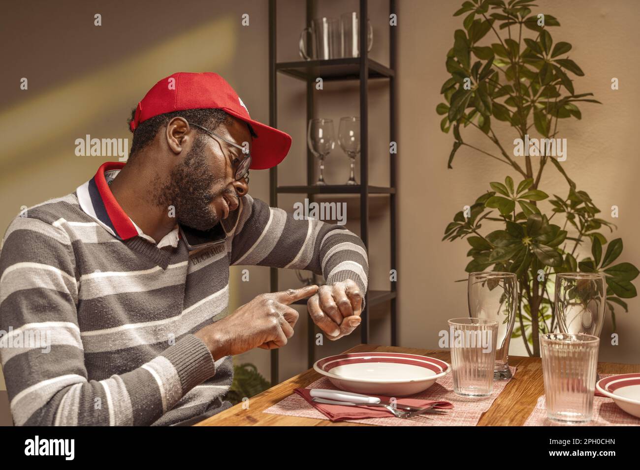 Young African man in casual clothes and red backwards hat, sitting at a restaurant table set for two with an empty chair, talking on the phone and poi Stock Photo