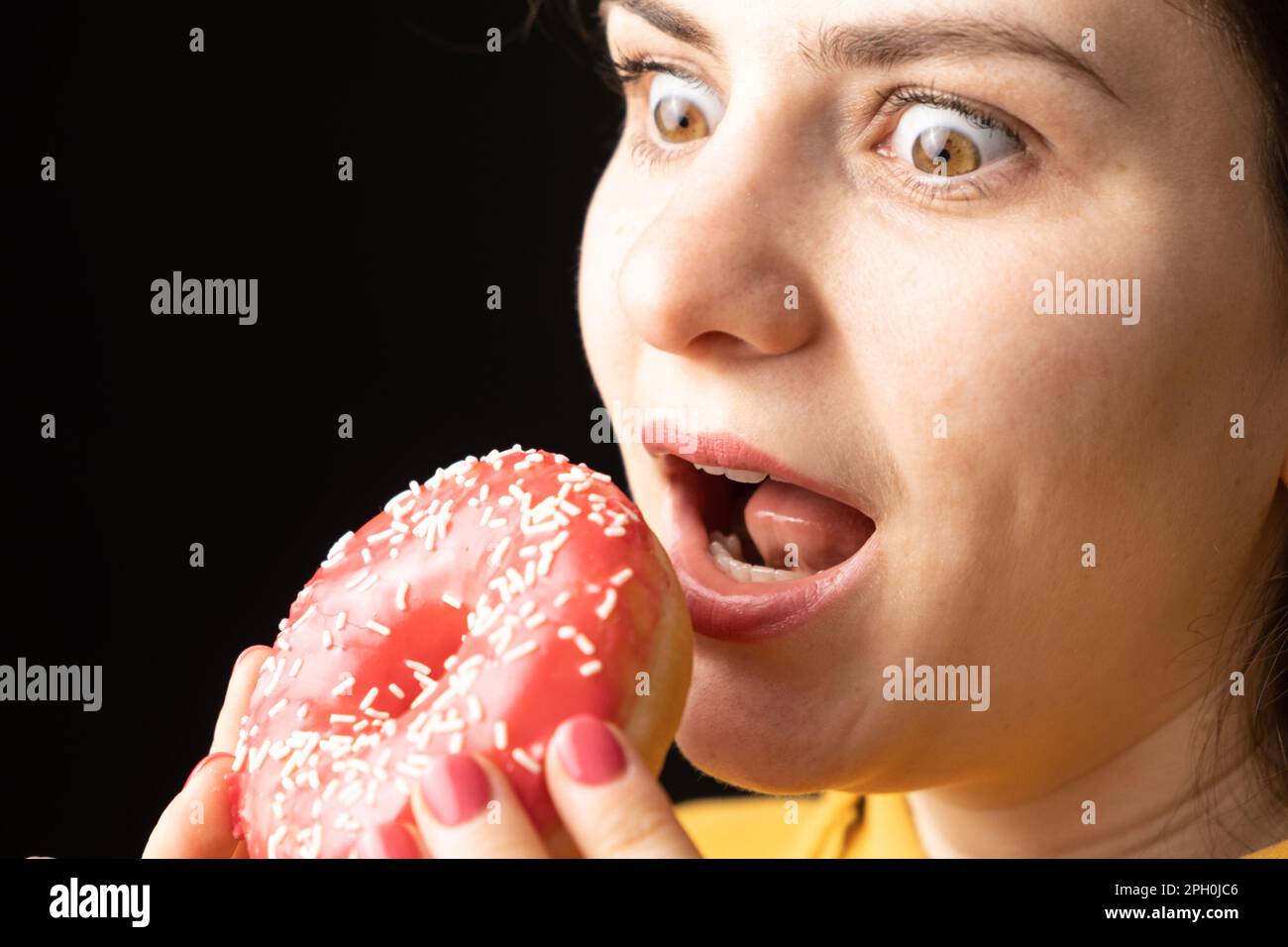 A woman bites a large red donut, a black background, a place for text. Gluttony, overeating and sugar addict Stock Photo