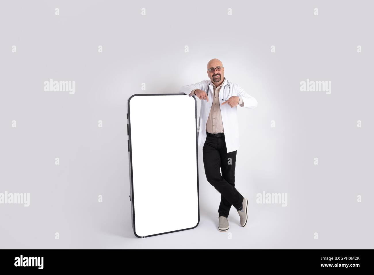 Bald underweight male doctor leaning big phone. Full body length image of middle aged bald physician pointing empty screen smartphone, mock up. Stock Photo