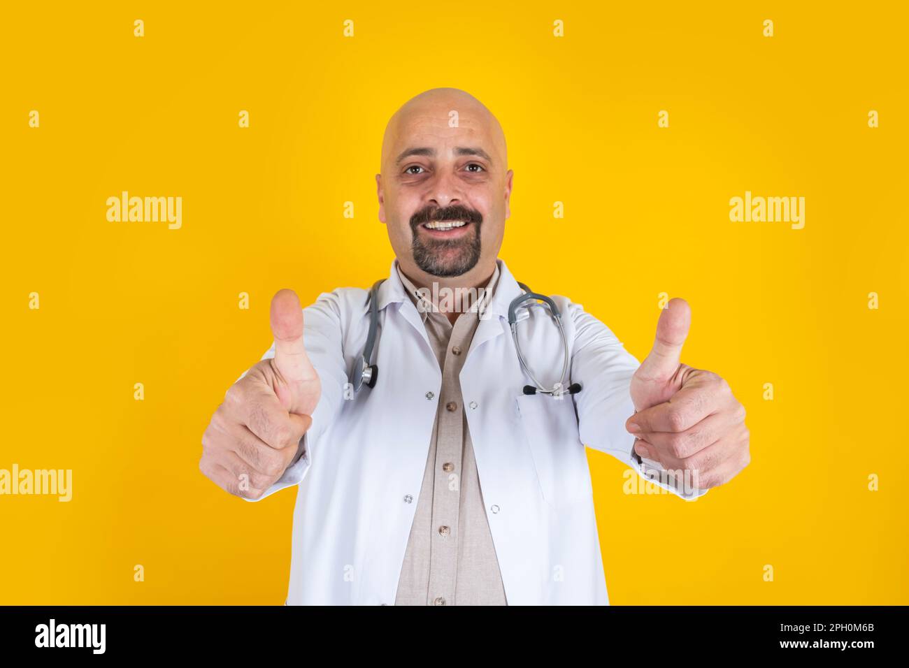 caucasian middle aged doctor happy thumbs up. Isolated yellow studio background. Looking camera, approving, successful treatment concept idea. Stock Photo