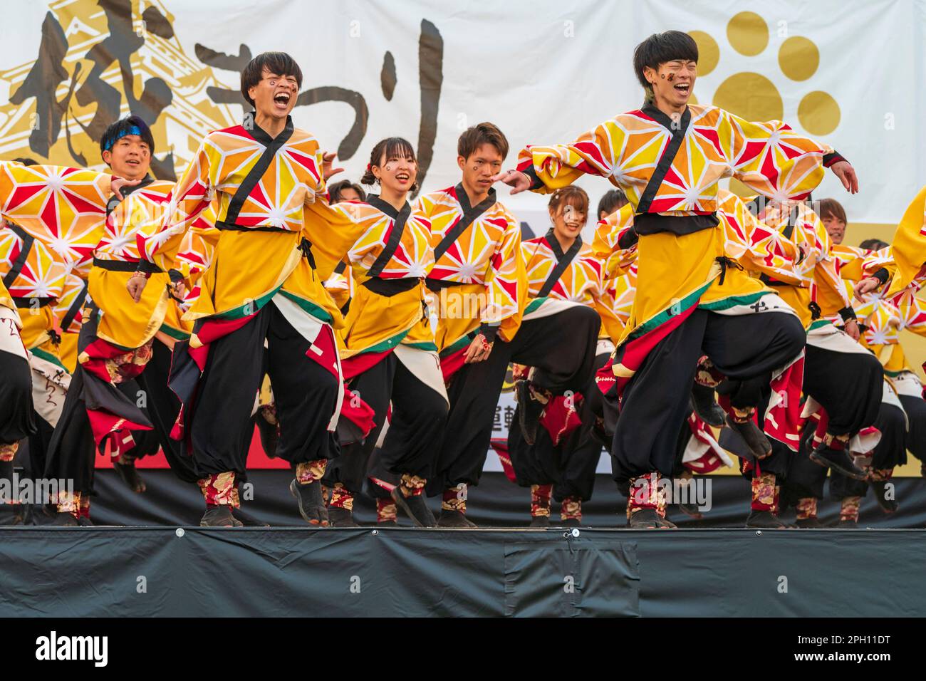 Mixed sex team of young adult Japanese dancers wearing orange yukata tunics  and black trousers on an outdoor stage performing Yosakoi dancing at the  annual March Kumamoto Kyusyu gassai dance festival Stock
