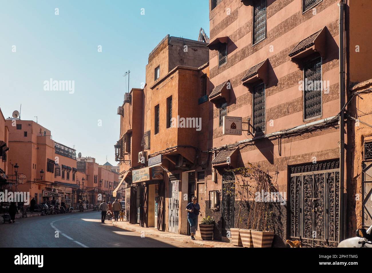Marrakech, Morocco - January 02, 2020: People walking in typical Moroccan city, sun shines to houses with decorated windows and gates, walls made of r Stock Photo