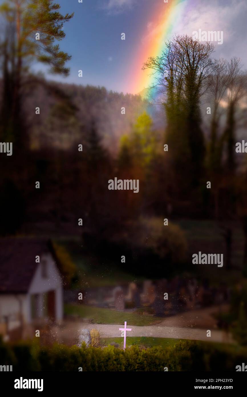 Cross in bright light by cemetery with colourful rainbow. Concept of hope during mourning. Live after death metaphor, spirituality. Stock Photo
