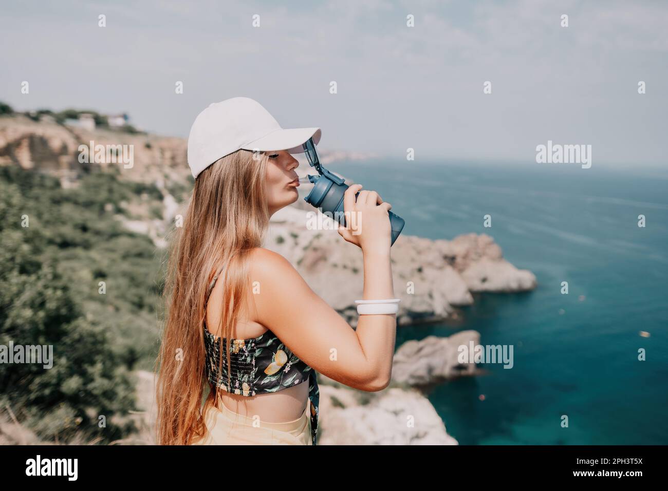 Woman travel sea. Happy tourist drink water on hot summer day. Woman traveler looks at the edge of the cliff on the sea bay of mountains, sharing Stock Photo