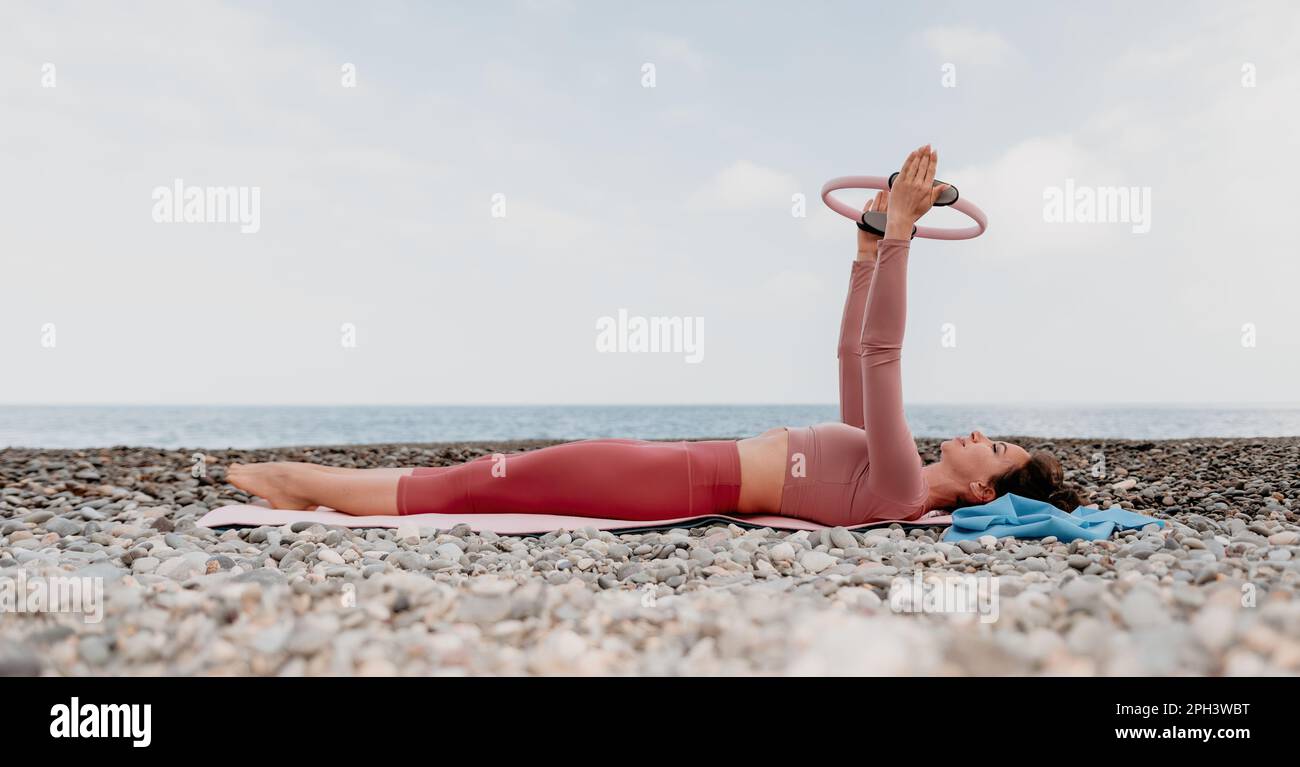 Woman sea pilates. Sporty happy middle aged woman practicing fitness on beach near sea, smiling active female training with ring on yoga mat outside Stock Photo