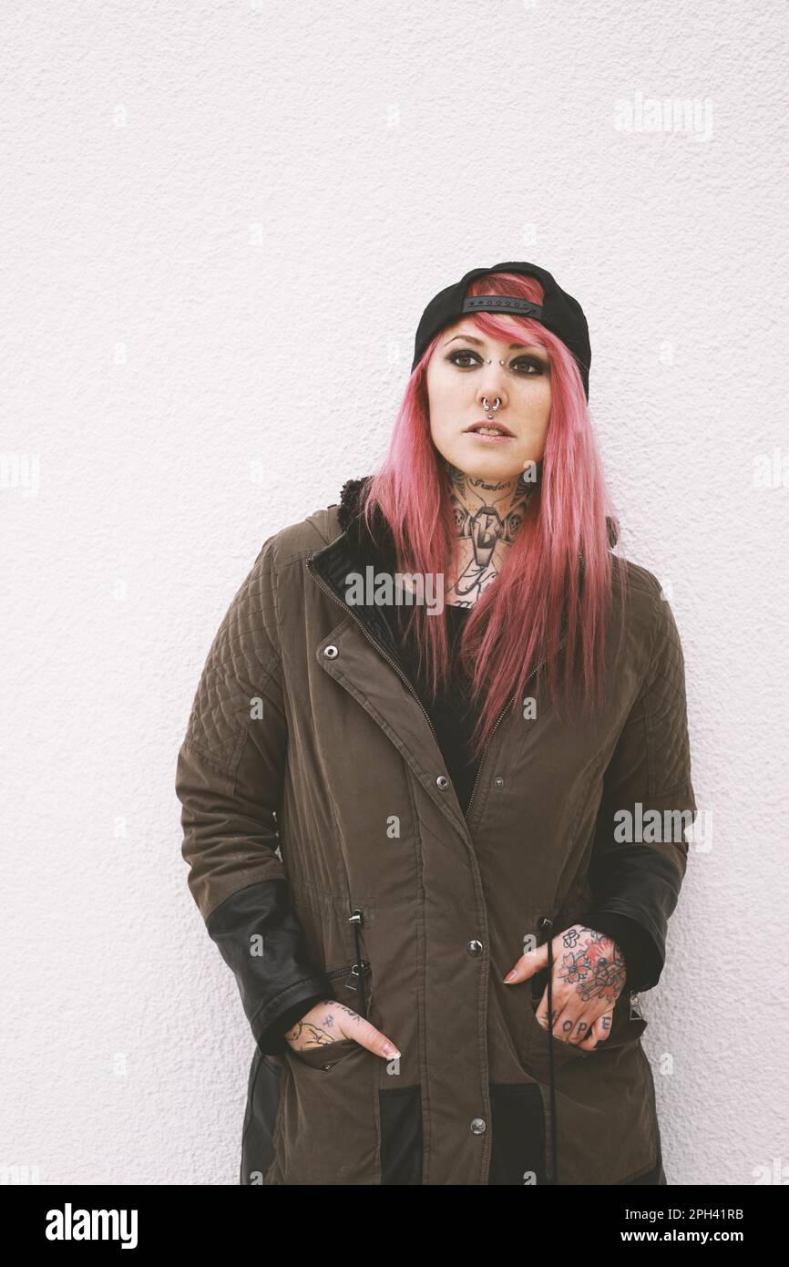 young woman with pink hair piercings and tattoos wearing parka and baseball cap leaning against wall Stock Photo