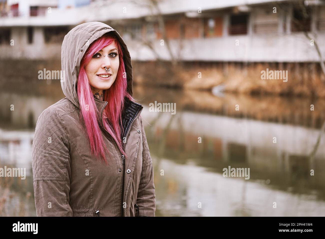 pierced young woman with pink hair and hooded parka standing by the river Stock Photo