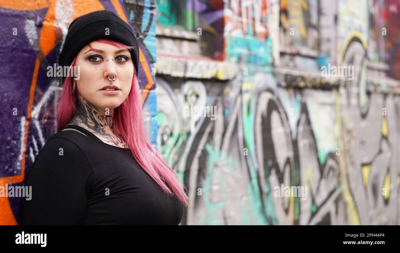 young urban woman with pink hair piercings and tattoos leaning against grafitti wall - real people alternative lifestyle Stock Photo