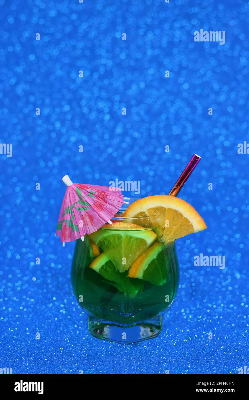 A cold, tropical, green coloured drink in a glass with orange slices, lime and parasol on a vibrant, bright, sparkly blue background Stock Photo