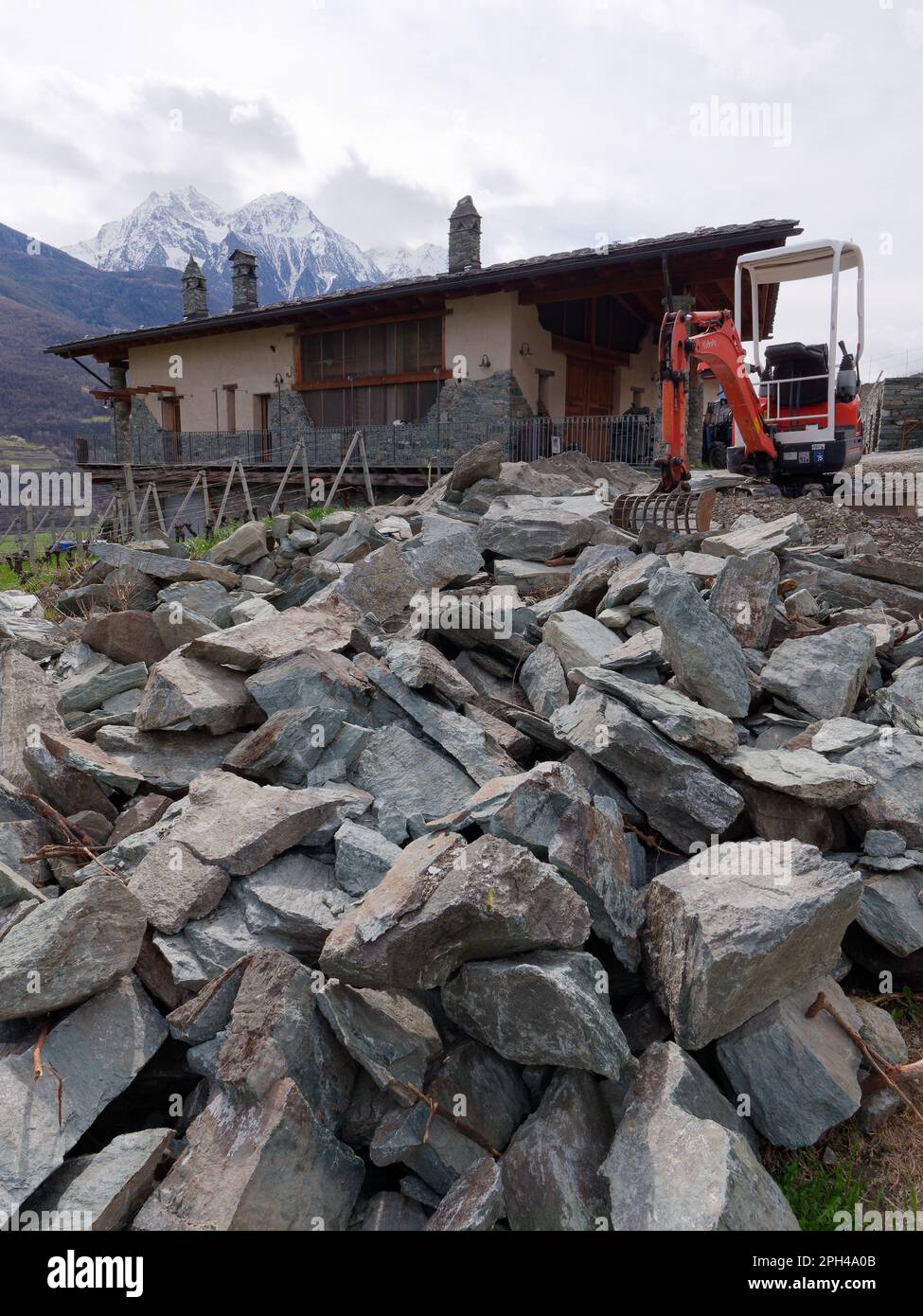 Les Grange farmhouse in Nus in the Aosta Valley Italy with large pieces of stone in front and a digger. Snow capped mountains behind Stock Photo