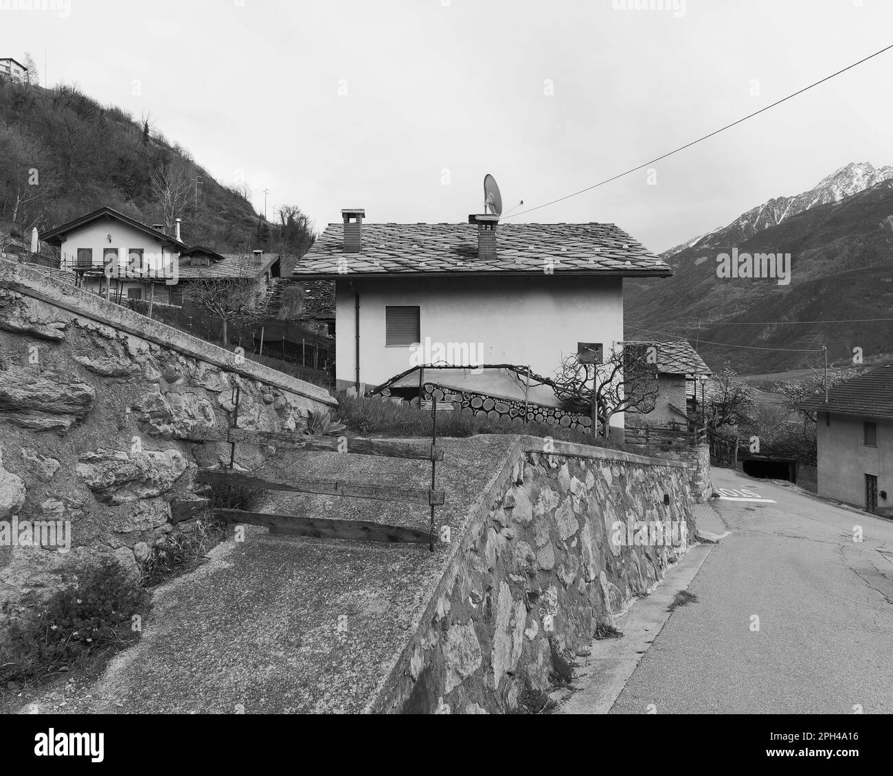 Quaint house with wood stacked. Stone walls and a ramp mark the boundary from the road. Nus, Aosta Valley, Italy. Monochrome Stock Photo