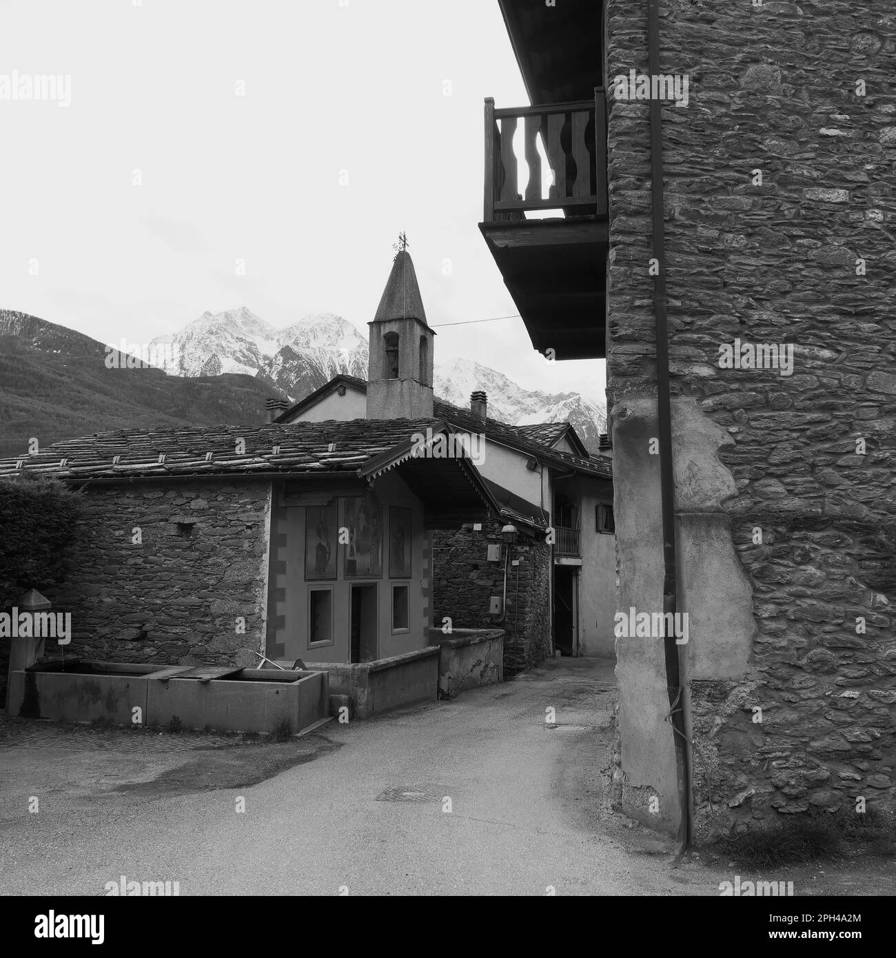 Village in Nus with a church and house with balcony. Aosta Valley, Italy. Water trough foreground and snow capped alps behind. Monochrome Stock Photo