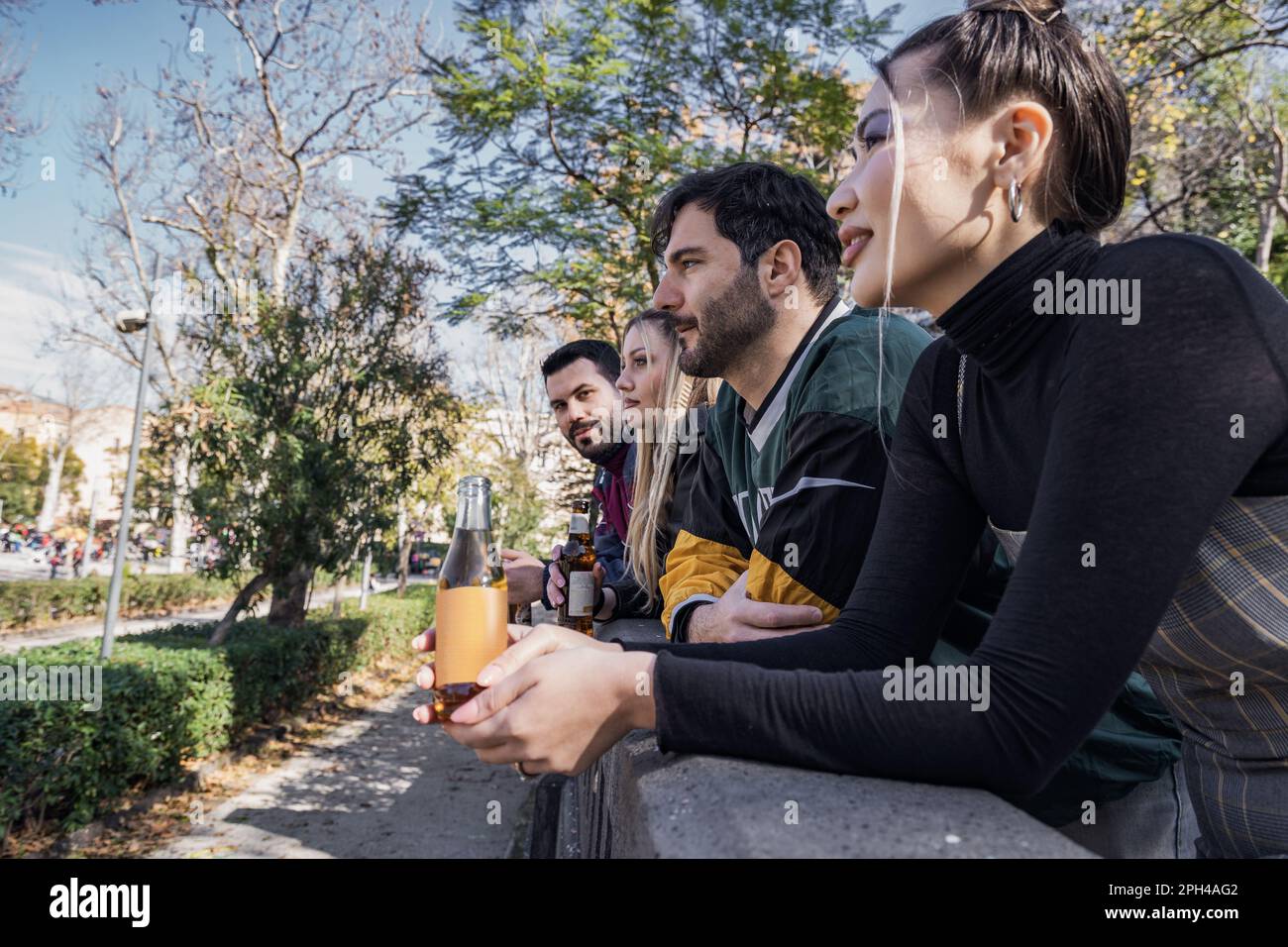 Two men and two women relax together, leaning on a stone railing, holding bottled drinks (beer and soda). Profile view, diverse group with Caucasian a Stock Photo