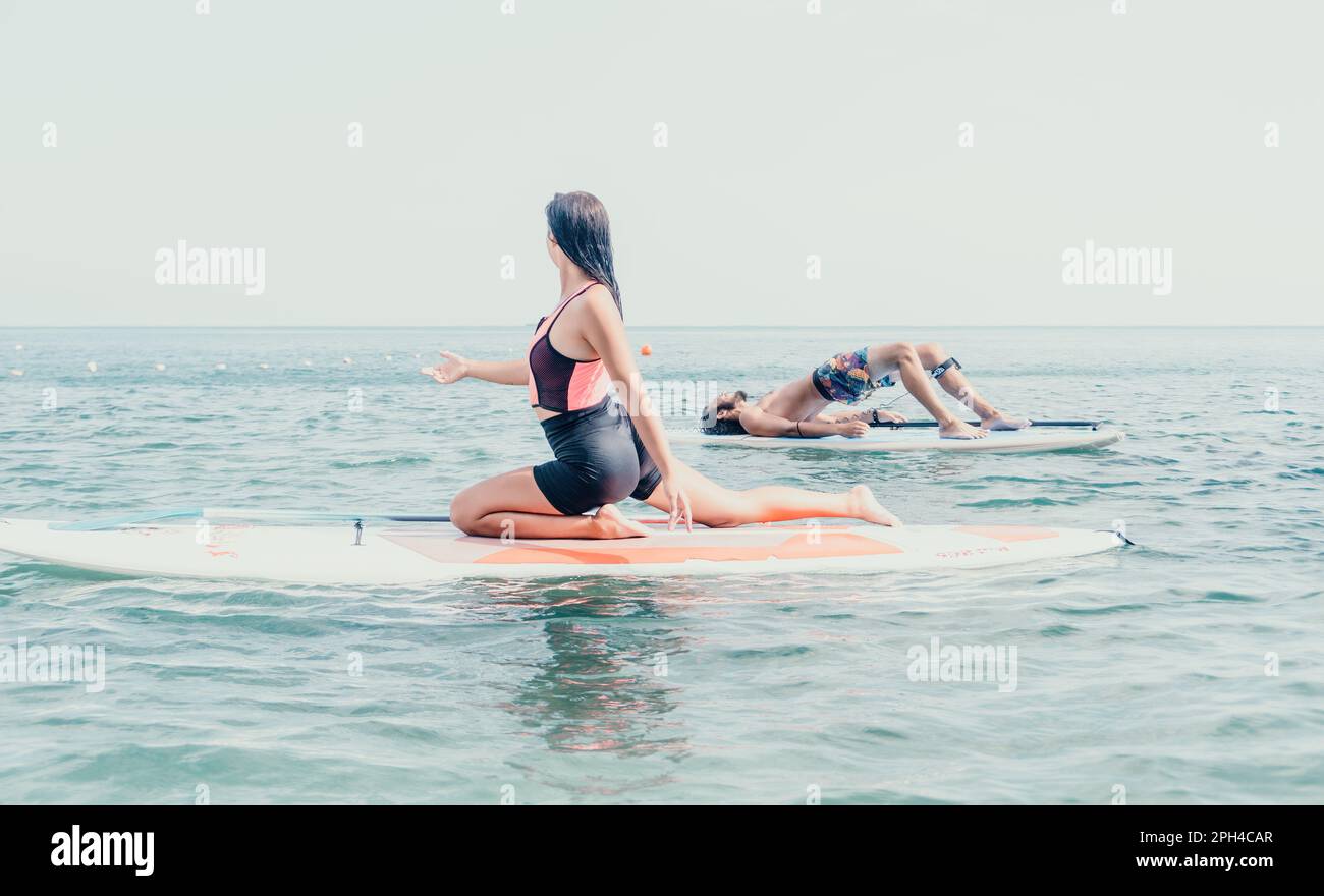 Woman man sup yoga. Happy young sporty couple practising yoga pilates on paddle sup surfboard. Female stretching doing workout on sea water. Modern Stock Photo