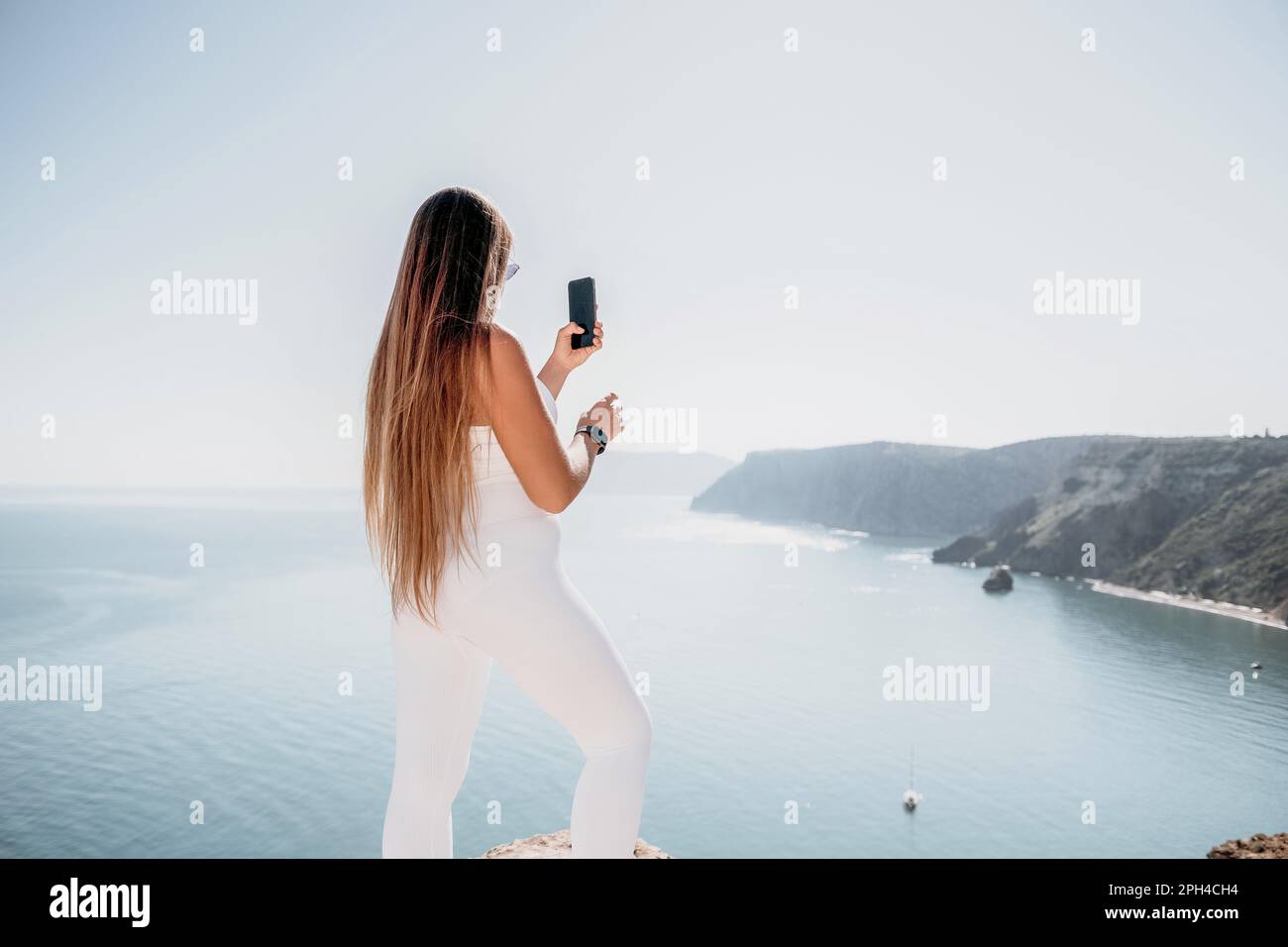Digital nomad, woman in the hat, a business woman with a laptop sits on the rocks by the sea during sunset, makes a business transaction online from a Stock Photo