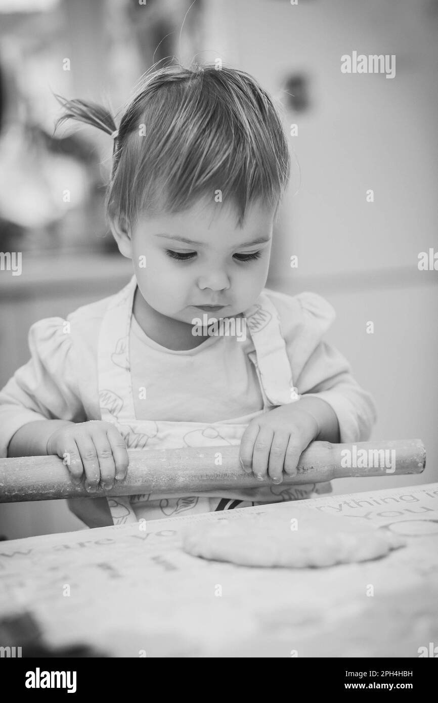Adorable baby in an apron is rolling out the dough in the kitchen Stock