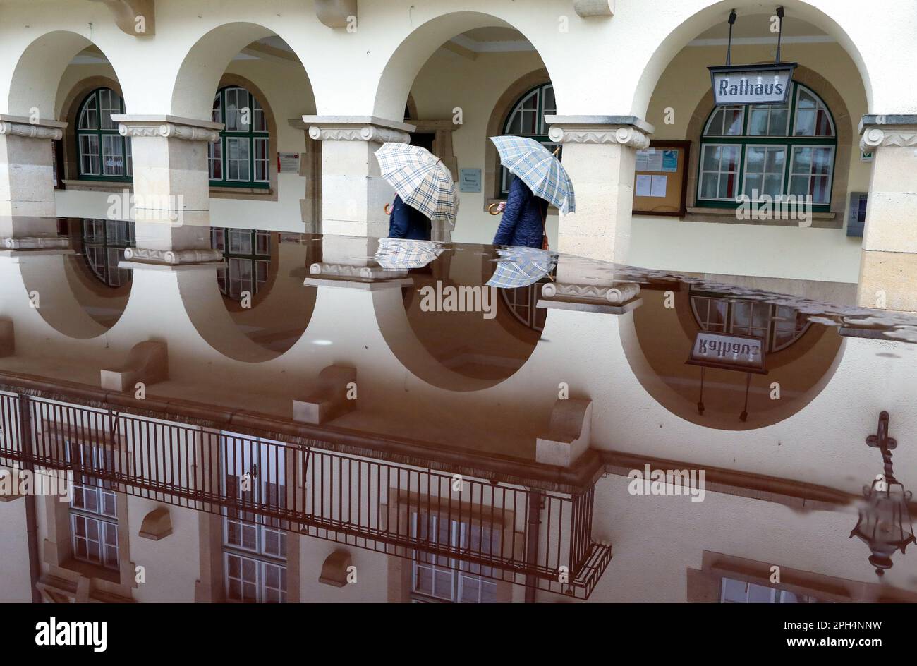 Sigmaringen, Germany. 26th Mar, 2023. The town hall of Sigmaringen is reflected in a puddle of rain, and two passers-by with umbrellas are just passing by. Credit: Thomas Warnack/dpa/Alamy Live News Stock Photo