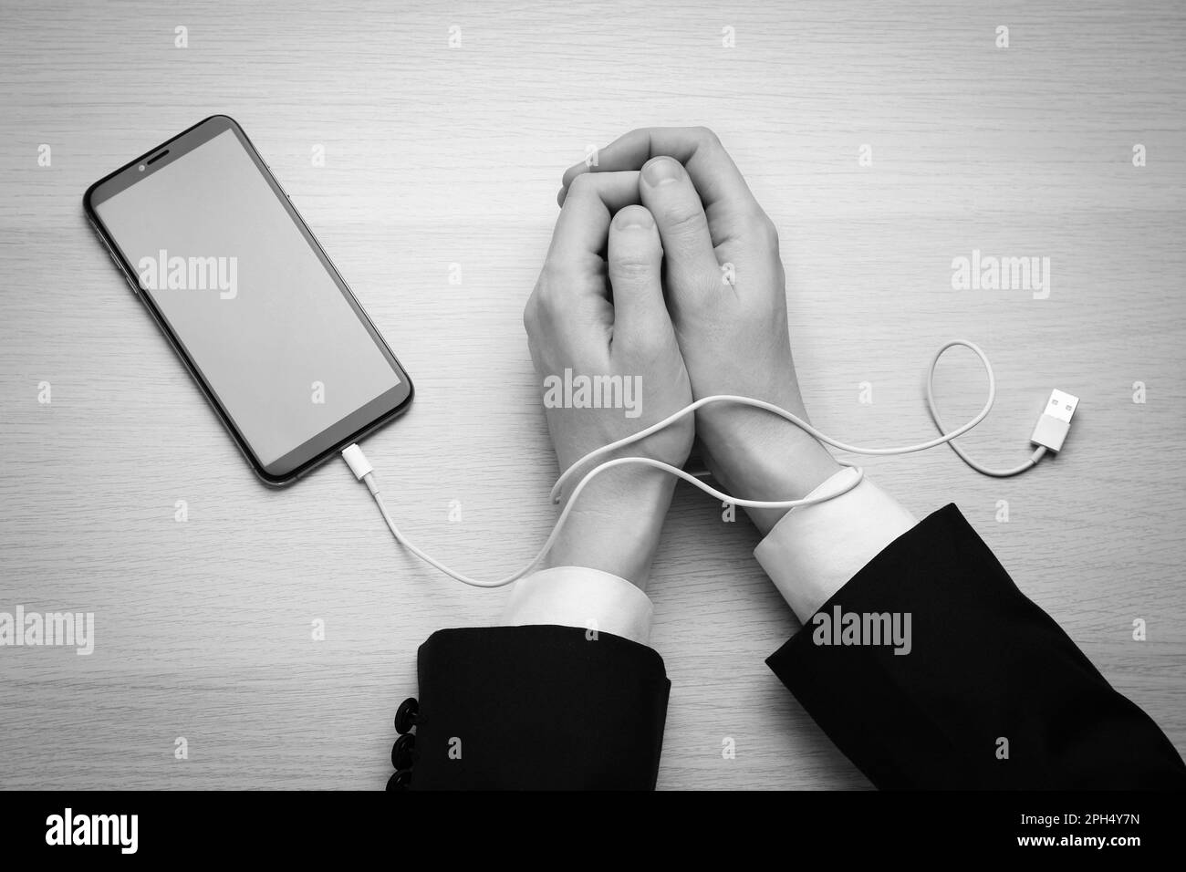 Top view of woman with her hands tangled in charging cable of smartphone at wooden table, black and white effect. Internet addiction Stock Photo