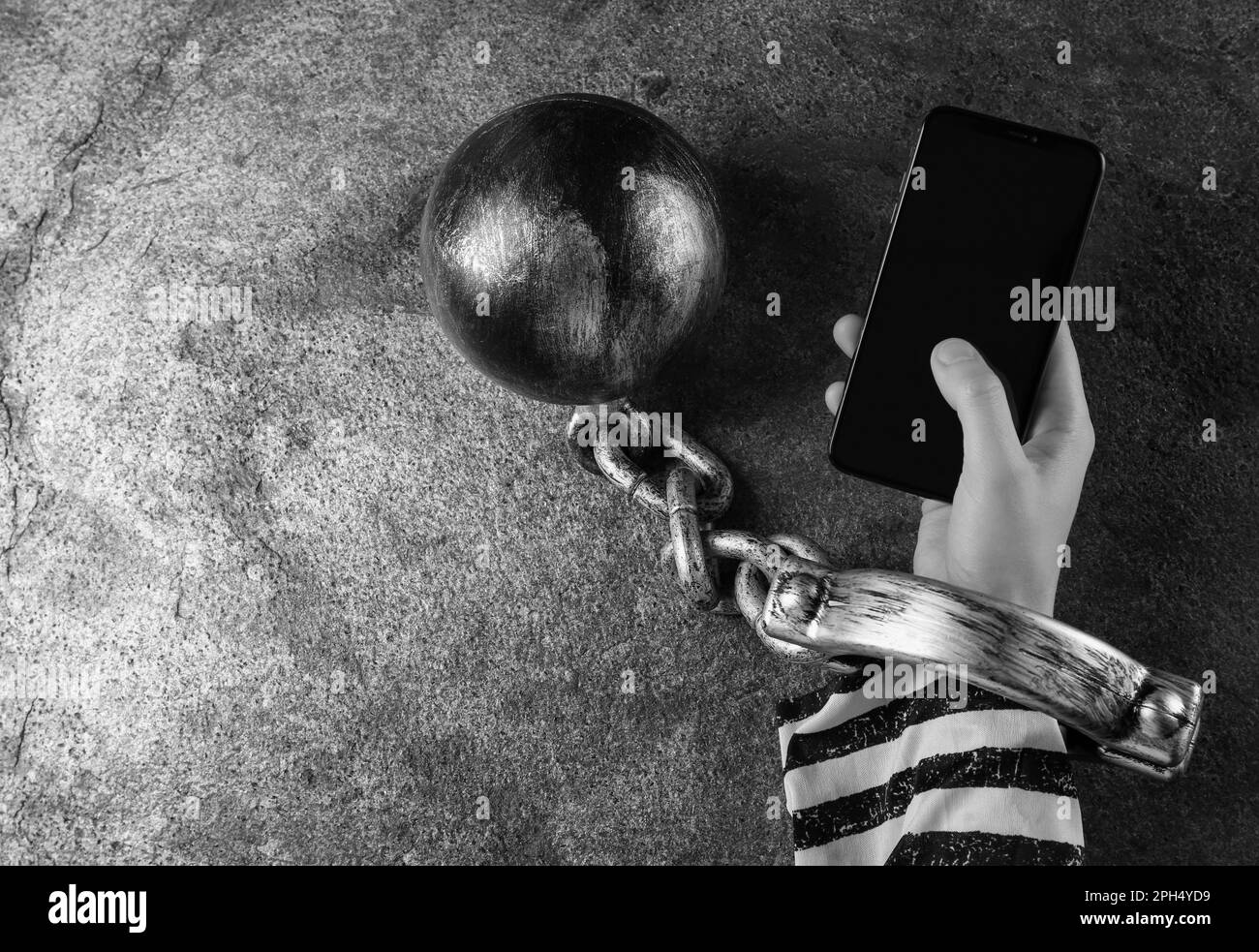 Top view of internet addicted woman in prisoner's clothes with ball and chain on her hand using smartphone at grey table, space for text. Black and wh Stock Photo