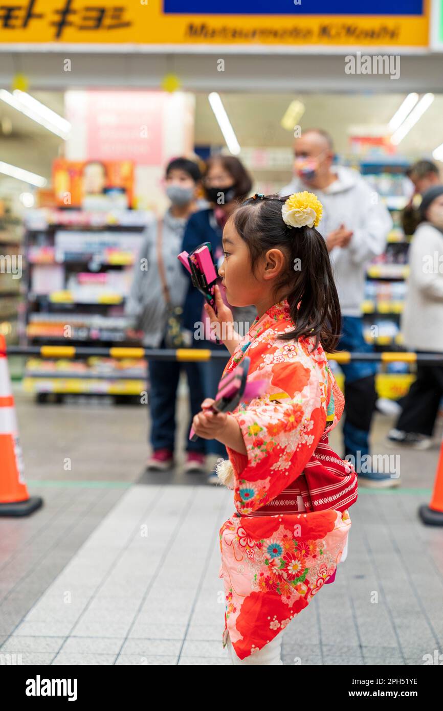 Young child yosakoi dancer dancing in a shopping street in Kumamoto during the annual Kyusyu gassai dance festival. Side view of the little girl dressed in red flower patterned yukata jacket. She holds pink and black naruko, clappers. Stock Photo